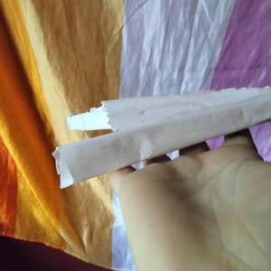 May include: A hand holding a rolled-up piece of paper against a colorful, striped background. The paper is light brown and white, with a torn edge. The background features orange, yellow, white, and purple stripes.