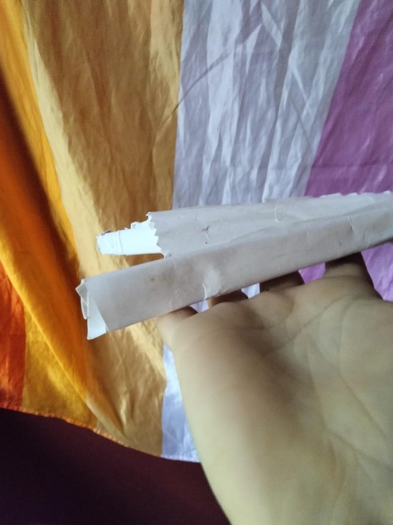 May include: A hand holding a rolled-up piece of paper against a colorful, striped background. The paper is light brown and white, with a torn edge. The background features orange, yellow, white, and purple stripes.