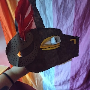 May include: A handmade brown leather mask with a red feather attached. The mask has a cut-out eye and is decorated with yellow and white accents. The mask is held up against a colorful background.