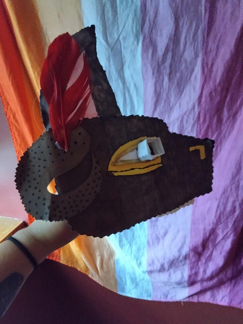 May include: A handmade brown leather mask with a red feather attached. The mask has a cut-out eye and is decorated with yellow and white accents. The mask is held up against a colorful background.