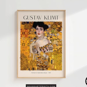 May include: A framed print of Gustav Klimt's painting "Portrait of Adele Bloch-Bauer" featuring a woman in a gold dress with a gold background. The painting is in a gold frame with the artist's name and the title of the painting printed below the image.