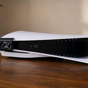 Peut inclure: Une console de jeu blanche et noire au design extérieur incurvé et aérodynamique. La façade comprend une section centrale noire brillante avec deux ports visibles et un cache noir à texture géométrique sur le bord gauche.