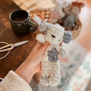 May include: A handmade, off-white crochet dog toy with gray accents on the ears and body. The plush toy is held in a person's hand. Other similar toys are in a basket in the background. A dark green mug and scissors are on a wooden surface.