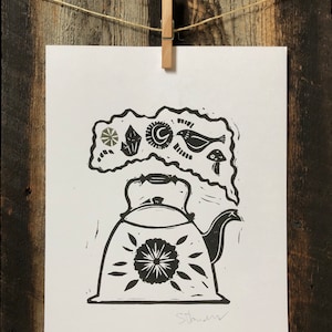 May include: Black and white linocut print of a teapot with steam rising from the spout. The steam is shaped like a cloud with various symbols and designs inside, including a crescent moon, a crystal, a bird, and a mushroom.