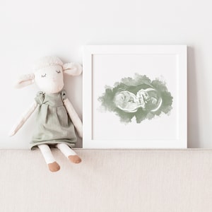 May include: A white frame containing a green watercolour-style ultrasound image of a baby. A plush white lamb doll wearing a green dress sits on a beige surface in front of the frame.