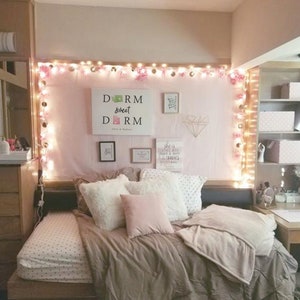 May include: A dorm room with a pink and white bedspread, a white headboard with string lights, and a sign that says "Dorm Sweet Dorm".