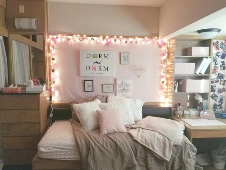 May include: A dorm room with a pink and white bedspread, a white headboard with string lights, and a sign that says "Dorm Sweet Dorm".