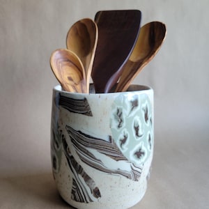 Puede incluir: Un porta utensilios cilíndrico de cerámica con base color crema, que presenta vetas marmoladas de color marrón oscuro y patrones circulares de color verde claro. Dentro hay cuatro utensilios de cocina de madera, incluyendo tres cucharas de color marrón claro y una espátula de color marrón oscuro.