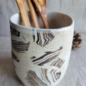 Puede incluir: Un porta utensilios cilíndrico de cerámica con un esmalte color crema moteado que presenta patrones abstractos de remolinos marrones y negros. Tres cucharas de madera con vetas visibles están colocadas dentro del recipiente.