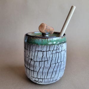May include: A handmade ceramic tumbler with a lid and a wooden straw. The tumbler has a textured, cracked white and gray exterior, a green rim, and a wooden knob on the lid. The straw is light-colored.