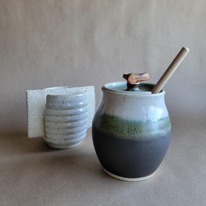 May include: A ceramic honey pot with a lid featuring a small wooden handle and a wooden dipper, displaying a gradient of white, green, and dark gray glaze. Beside it sits a white, ribbed ceramic sponge holder containing a rectangular sponge.