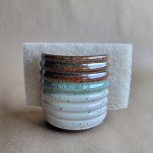 Puede incluir: Un soporte de cerámica para esponja con cuerpo cilíndrico y acanalado, base blanca moteada, sección central verde claro y borde esmaltado marrón oscuro. Una esponja abrasiva rectangular de color blanco roto se coloca verticalmente.