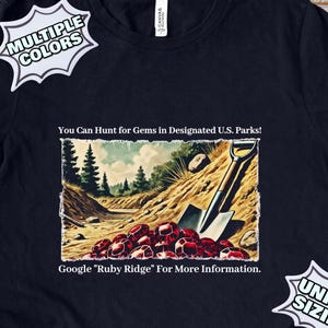 Può includere: Una maglietta blu scuro con testo bianco che recita "You Can Hunt for Gems in Designated U.S. Parks!" e "Google "Ruby Ridge" For More Information." La maglietta presenta un'illustrazione vintage di una pala e gemme rosse in un ambiente boschivo.