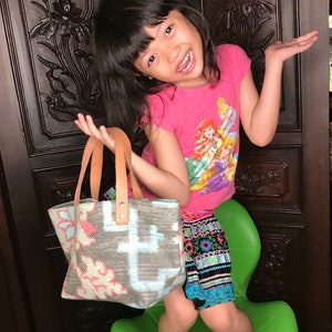 May include: A young person wearing a pink shirt with a cartoon print of princesses and a colourful skirt is holding a small, grey and white woven tote bag with leather handles. The bag has a floral pattern.