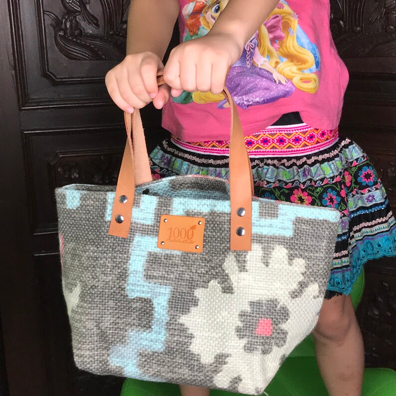 May include: A grey and blue woven tote bag with a brown leather handle and a brown leather tag that says "1000 handles".