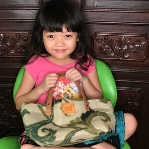 May include: A young girl wearing a pink shirt holds a beige and green fabric tote bag with a brown leather handle. The bag has a small rectangular tag with the word "1000" printed on it. The bag has a cartoon character design on the front.