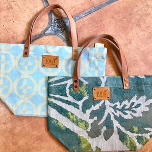 May include: Two canvas tote bags with leather handles and brown leather patches that say "1000 roads". One bag is blue and white with a geometric pattern. The other bag is green and white with a leafy pattern.