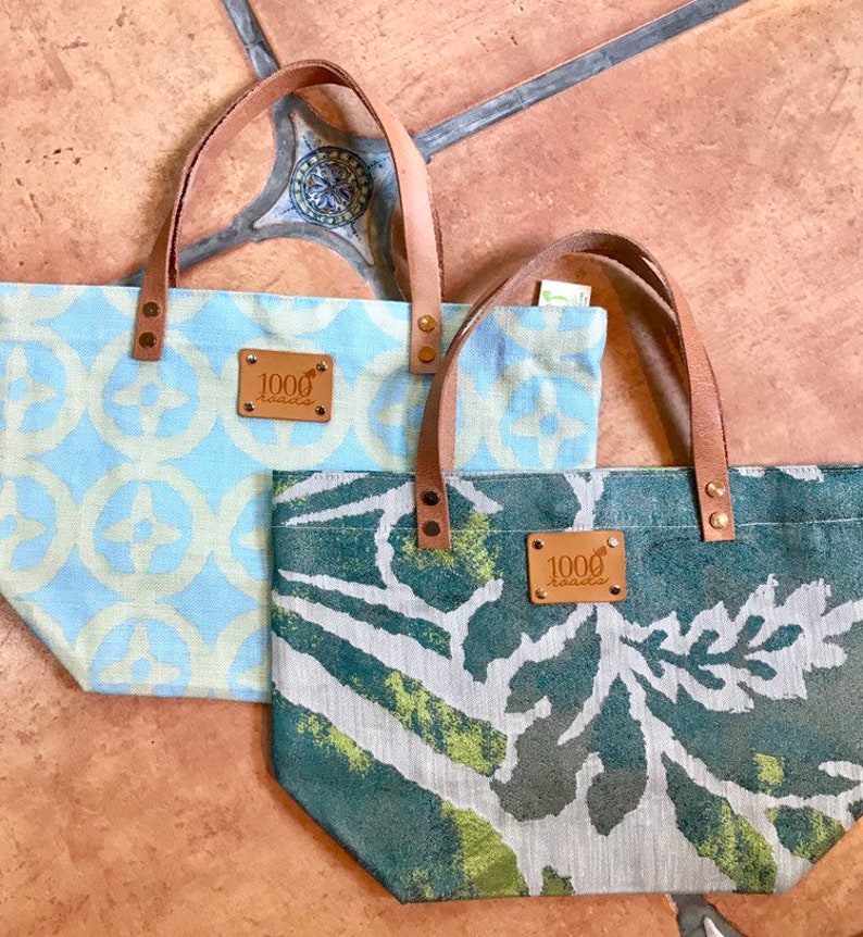 May include: Two canvas tote bags with leather handles and brown leather patches that say "1000 roads". One bag is blue and white with a geometric pattern. The other bag is green and white with a leafy pattern.