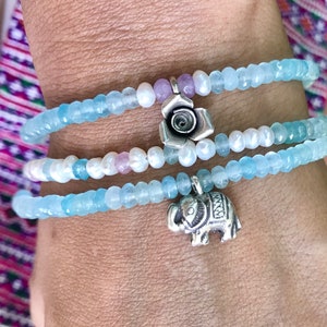 Può includere: Tre braccialetti di perline con perline di acquamarina e perle. Un braccialetto ha un ciondolo a forma di rosa argentata e un altro ha un ciondolo a forma di elefante argentato. I braccialetti sono avvolti attorno a un polso.