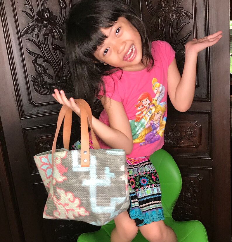 May include: A young person wearing a pink shirt with a cartoon print of princesses and a colourful skirt is holding a small, grey and white woven tote bag with leather handles. The bag has a floral pattern.