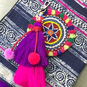 Può includere: Un portachiavi con nappa colorata a forma di pompon con un ciondolo rotondo di perline. Il ciondolo ha un design multicolore con una stella al centro. La nappa è viola e rosa con un cordoncino rosso.