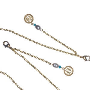 Può includere: Collana a catena dorata con due ciondoli, ognuno con un designo di nodo cinese dorato e una perla bianca. La collana ha una chiusura a moschettone.