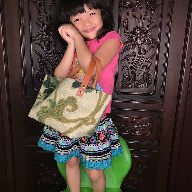 May include: A young girl wearing a pink shirt and a colourful skirt holds a green and white patterned tote bag with brown leather handles.