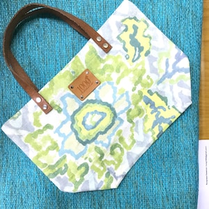 May include: A white tote bag with a green, blue and yellow floral pattern. The bag has brown leather handles and a brown leather tag with the text "1000 Hawaii" on it.