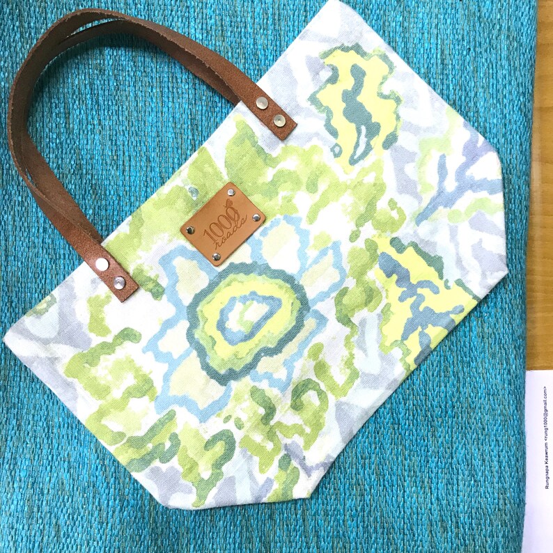 May include: A white tote bag with a green, blue and yellow floral pattern. The bag has brown leather handles and a brown leather tag with the text "1000 Hawaii" on it.