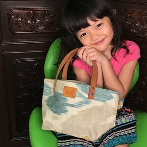 May include: A small, light blue and white patterned tote bag with brown leather handles and a brown leather tag that says "1000" on it. The bag is being held by a young girl wearing a pink shirt and a colourful skirt.