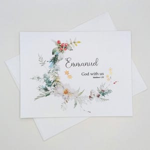 May include: A white greeting card featuring a watercolor wreath of green pine branches, red berries, and white flowers. The text reads Emmanuel God with us Matthew 1:23 in black script and serif fonts, accented by small gold star illustrations.