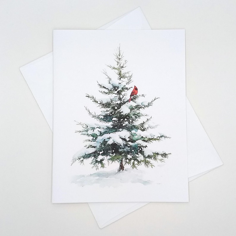 May include: A watercolor illustration of a snow-covered evergreen tree with a bright red cardinal perched on a branch. The image is on a white card with a matching envelope, evoking a winter or holiday theme.