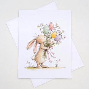 May include: A white greeting card featuring a watercolor illustration of a light brown rabbit holding a bouquet of colorful patterned Easter eggs and small white flowers tied with a pink ribbon, standing on a patch of green grass.