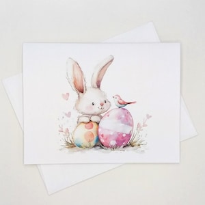May include: A rectangular greeting card featuring a watercolor illustration of a white bunny sitting behind two colorful Easter eggs. A small pink bird is perched on the larger pink egg. The design includes soft pink hearts and grass accents.