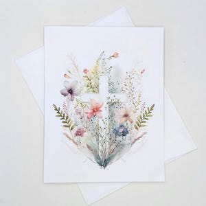 May include: A white greeting card with a watercolor illustration of a cross surrounded by colorful flowers and greenery. The card is accompanied by a white envelope, suitable for various occasions.