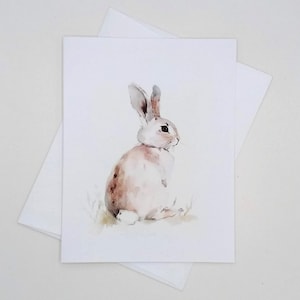 May include: Watercolor illustration of a rabbit on a greeting card. The rabbit is depicted in shades of white, brown, and pink, with long ears and a fluffy body. The card is paired with a white envelope, perfect for sending a note.