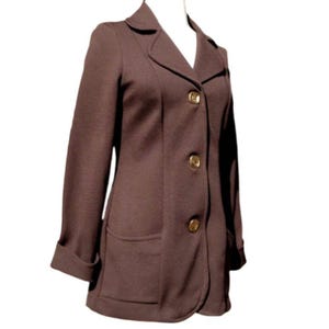 以下が含まれることがあります： ノッチカラー、2つのフロントポケット、3つの金色のボタンが付いた茶色のブレザー。袖口は折り返されています。ブレザーは白いマネキンに展示されています。