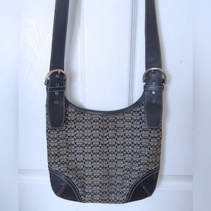 May include: A hobo-style shoulder bag featuring a dark gray and black repeating monogram pattern on the main body. The bag includes black leather trim, silver-tone metal buckles on the strap attachments, and white contrast stitching throughout.