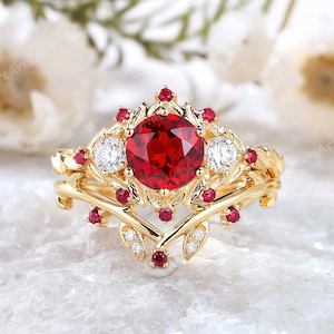 以下が含まれることがあります： 赤い大きな宝石を、小さな赤い宝石と白いダイヤモンドで囲んだ金の指輪。繊細な葉のデザインが施されています。
