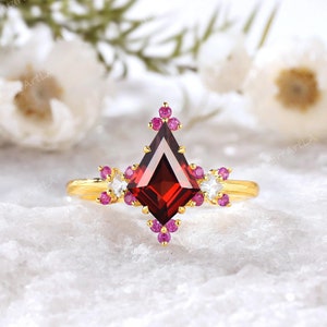 May include: A gold ring with a large, red, kite-shaped gemstone surrounded by smaller, pink gemstones. The ring is set on a white, textured surface.