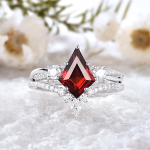 May include: A silver ring featuring a large, red, square-shaped gemstone surrounded by smaller, clear gemstones. The ring has a delicate, intricate design with a band that wraps around the finger twice.