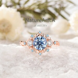 May include: A delicate rose gold engagement ring featuring a blue centre stone surrounded by smaller blue and white stones. The ring has a leafy design with a NatureArtLA Jewelry logo.