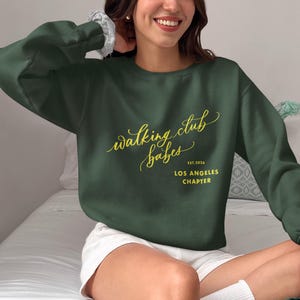 Puede incluir: Una sudadera verde oscuro con el texto "walking club babes" en letras doradas. El texto "EST. 2024 LOS ANGELES CHAPTER" también está en letras doradas debajo del texto principal.