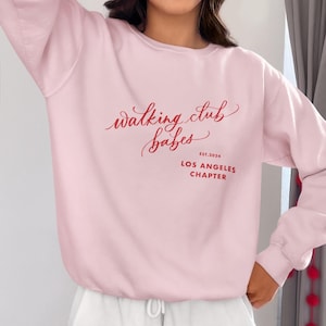 Puede incluir: Sudadera rosa con el texto "walking club babes est. 2024 Los Angeles Chapter" en rojo.