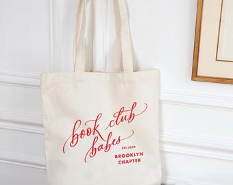 Pode incluir: Uma bolsa tote de lona branca com texto vermelho que diz "book club babes est. 2024 Brooklyn Chapter".