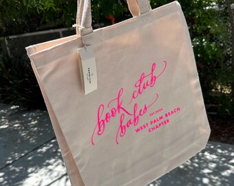 Pode incluir: Uma bolsa de lona bege com letras cor-de-rosa que diz "book club babes est. 2024 West Palm Beach Chapter".