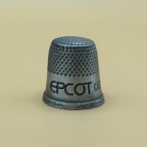 Puede incluir: Un dedal de plata con la palabra "EPCOT" grabada en el lateral.