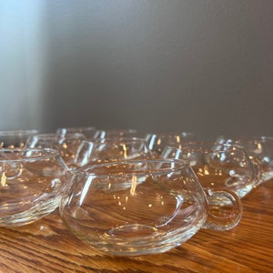 May include: A group of clear glass cups with rounded bottoms and handles. The cups are arranged in a staggered pattern on a wooden surface.