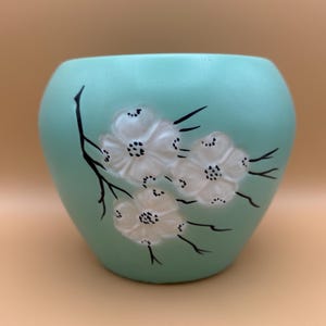 Op de afbeelding: Een afgeronde, licht mintgroene keramische plantenpot met een verhoogd, reliëf ontwerp van drie witte bloemen bevestigd aan dunne, zwart geschilderde takken. De bloemen hebben gedetailleerde centra met kleine zwarte stippen en een textuur.