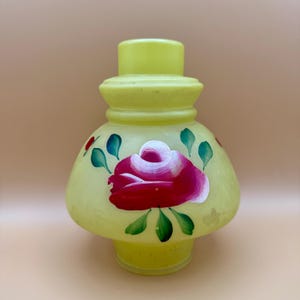 May include: A yellow glass lamp shade with a hand-painted red and white rose design. The shade has a rounded base and a cylindrical top. Green leaves and red accents surround the rose. The lamp shade is a vintage style.